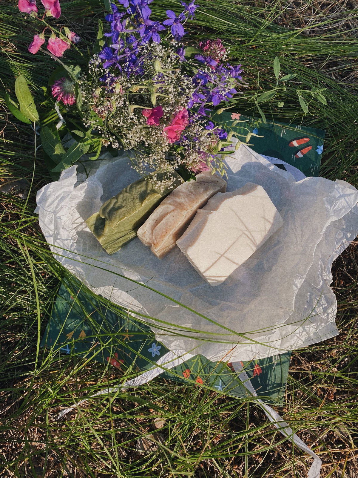 Three handmade bar soaps on parchment paper, set on grass with wildflowers